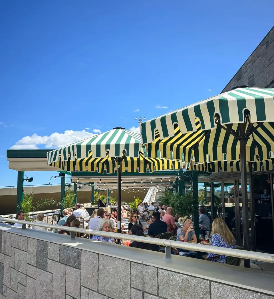 Gastronomieterrasse mit grün-weiß gestreiften Schirmen unter blauem Himmel, Gäste sitzen an den Tischen